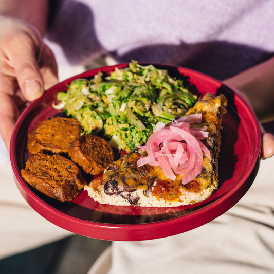 A close-up of a person holding a red plate with a slice of quiche, two browned patties, and a green slaw, garnished with pink pickled onions. The person is wearing a light purple top and cream pants.
